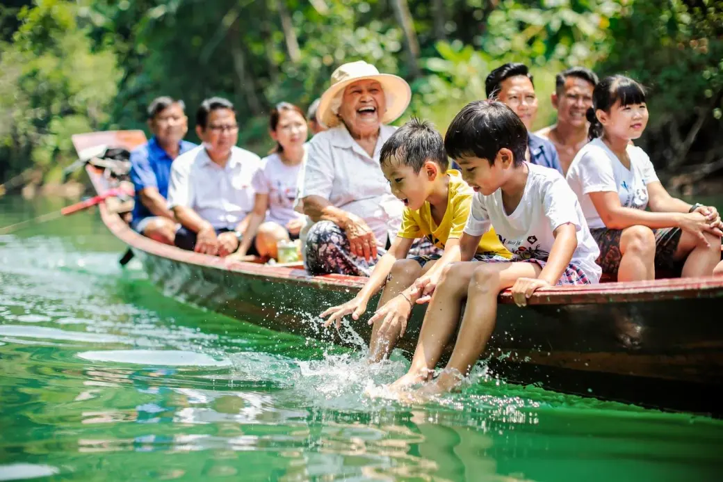 ครอบครัวนั่งเรือหางยาวบนแม่น้ำ เด็ก ๆ แหย่มือลงน้ำ คุณยายใส่หมวกกันแดดหัวเราะ